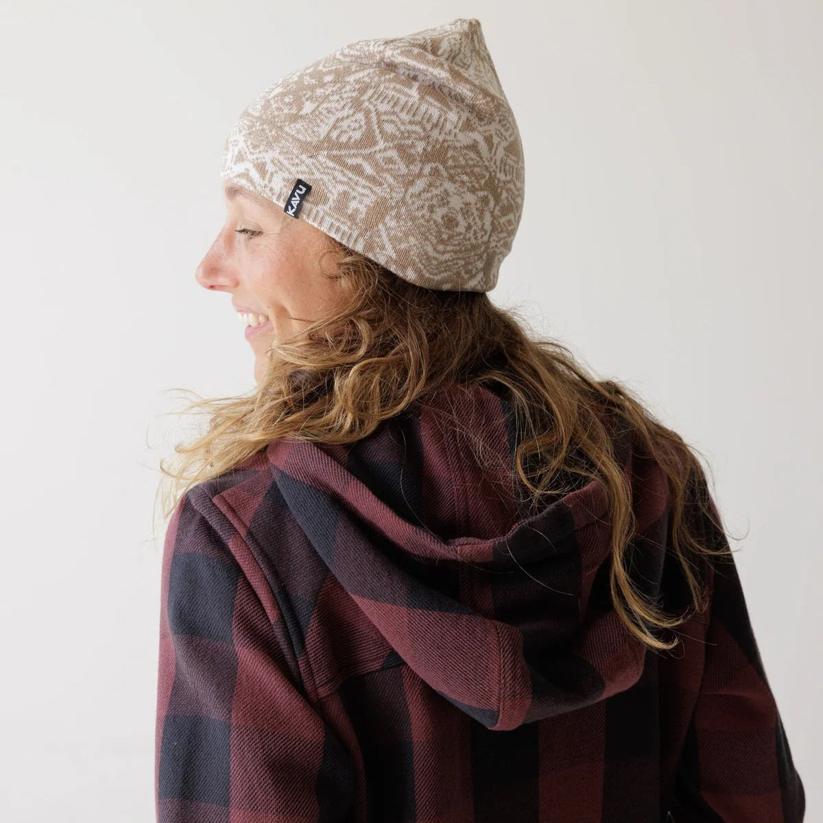 A woman shown from the side and back wearing a beige knit beanie with a white Nordic-style pattern and a dark red and black buffalo plaid hoodie. The beanie has a small black brand tag on the side.