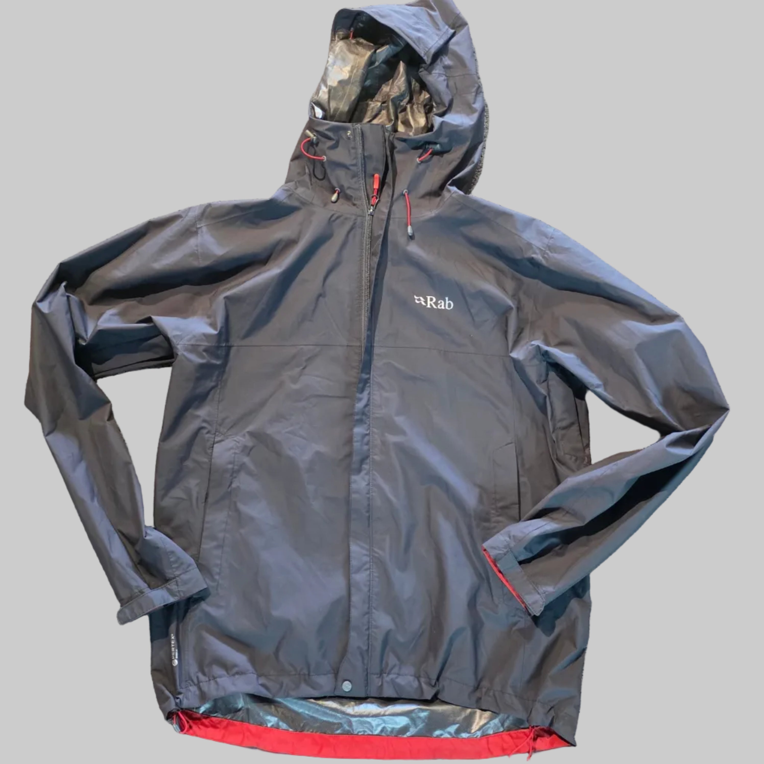A dark gray waterproof jacket laid flat on a textured beige carpet, showing a red inner lining on the left side and a small white Kipsta logo on the chest.