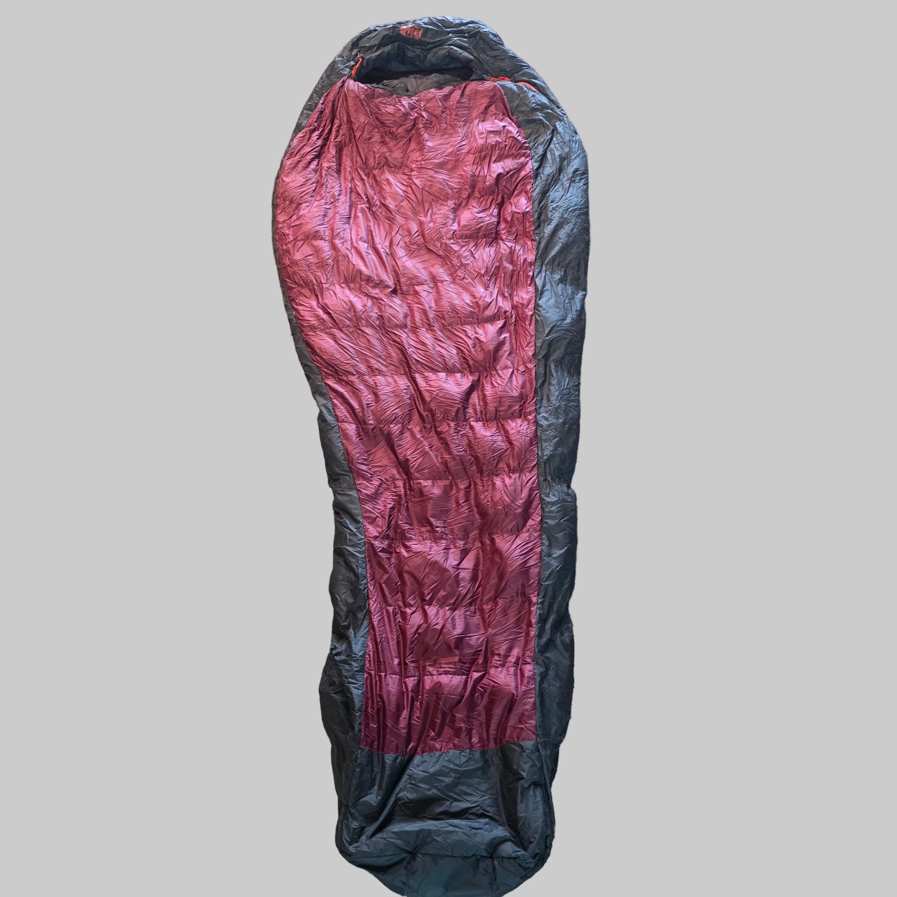 A mummy-style sleeping bag laid flat on a textured beige carpet, featuring a metallic rose-pink center panel and dark gray side panels.