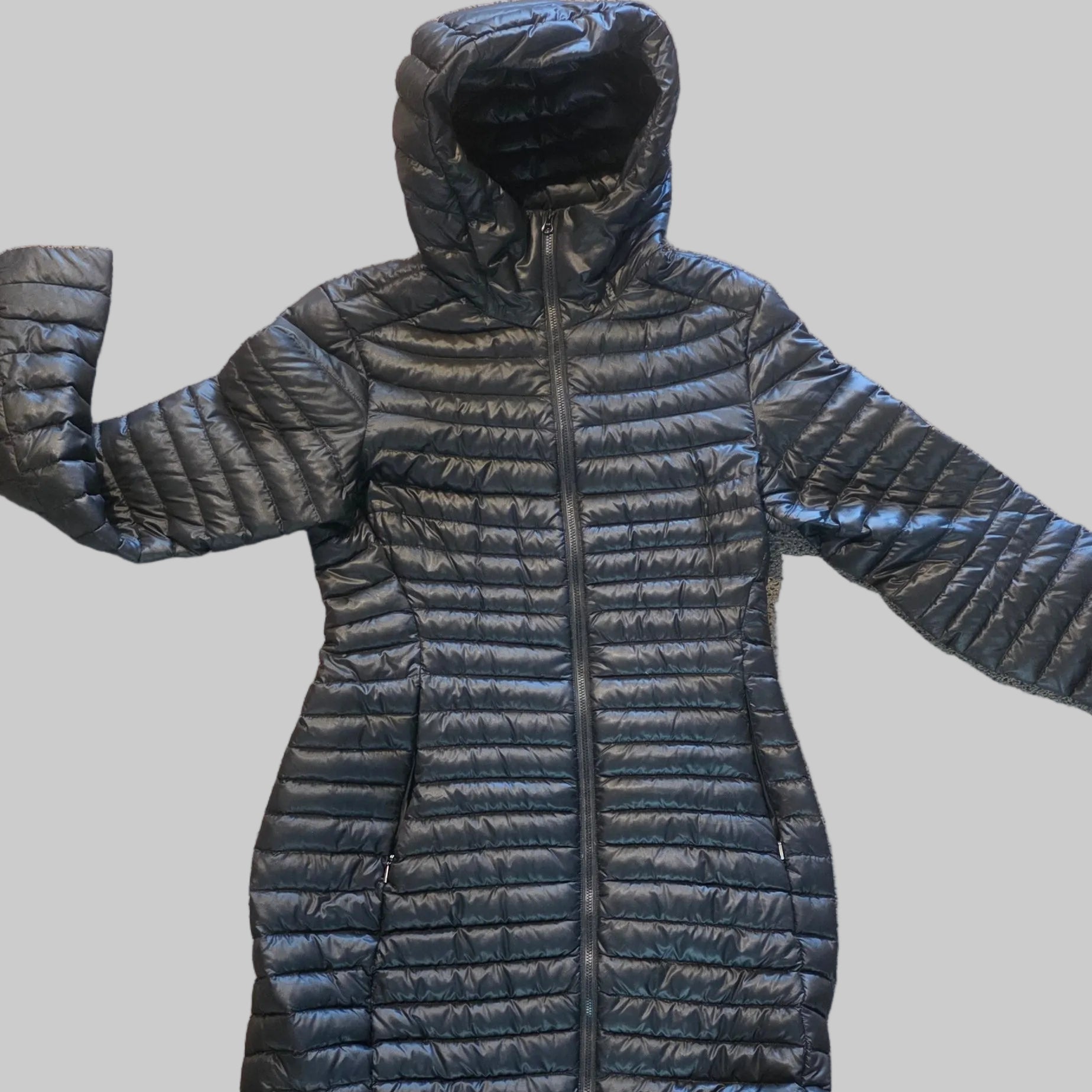 A black quilted puffer jacket with a hood and front zipper, laid flat on a beige carpeted floor. The feet of a person wearing sandals are visible at the bottom edge of the frame.