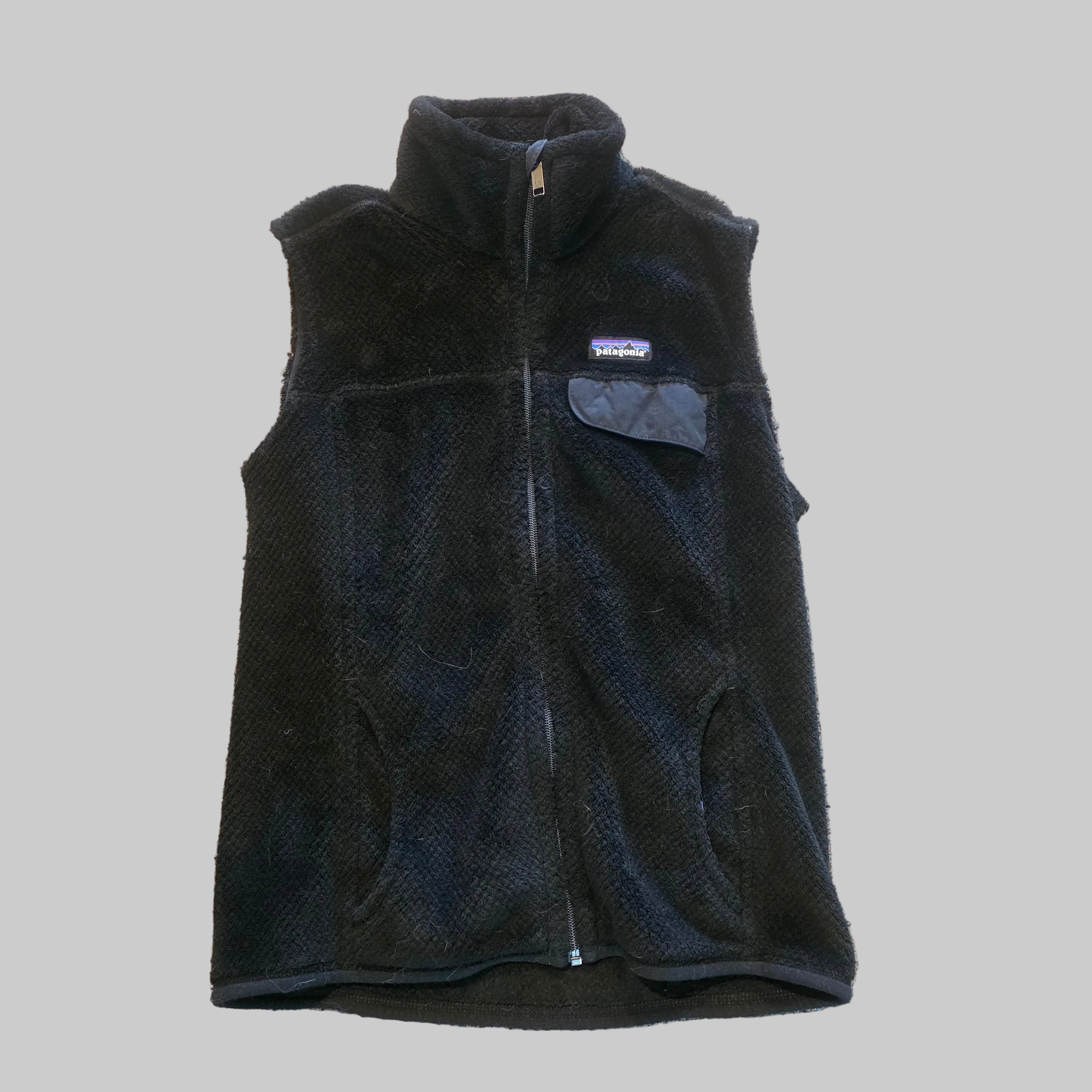 A black fleece vest laid flat on a textured beige carpet. The vest features a full-length front zipper, a stand-up collar, a chest pocket with a flap closure, and a small Patagonia logo patch on the left chest.