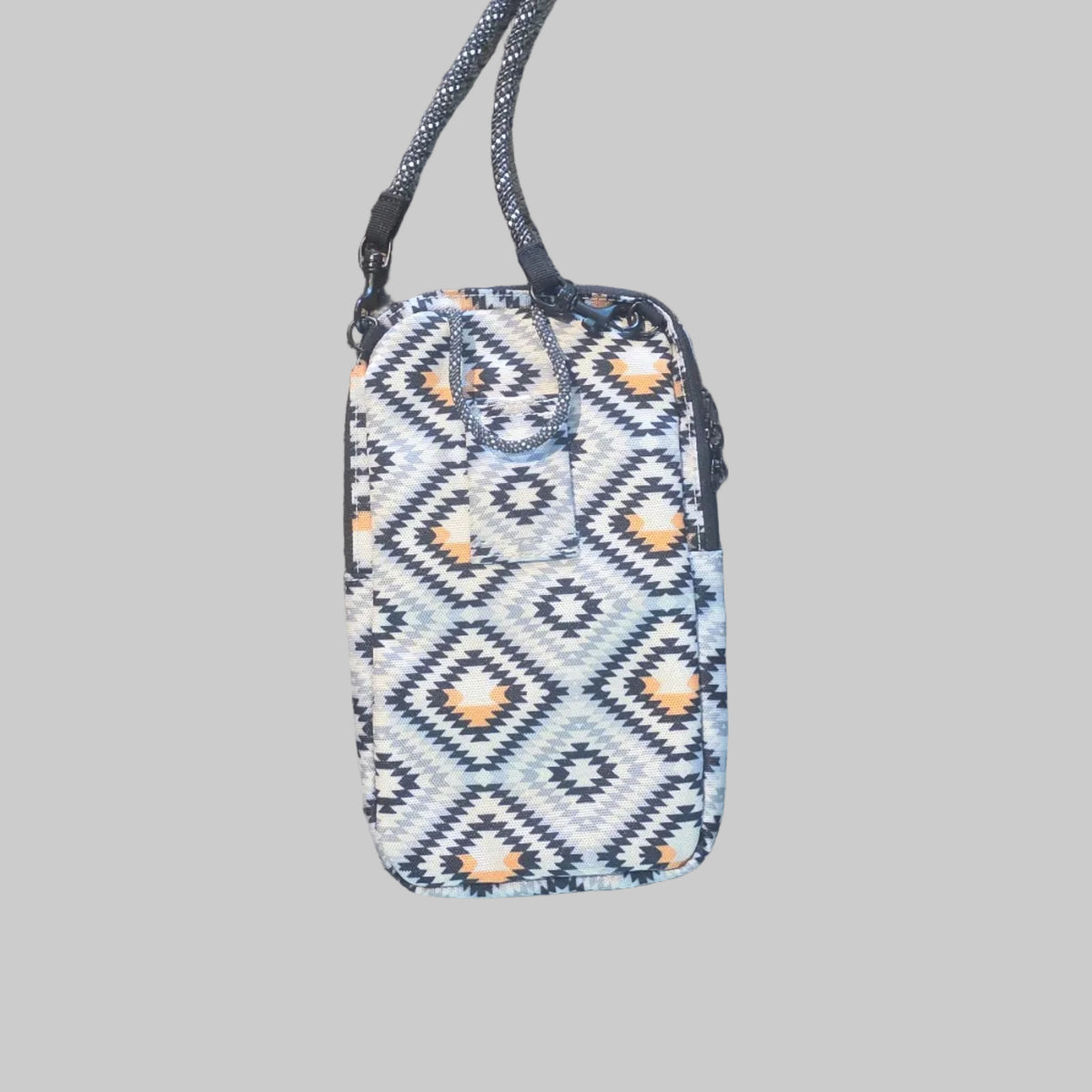 A small rectangular beaded pouch with a geometric diamond pattern in white, gray, and orange, featuring a matching gray beaded wrist strap, laid flat on a textured tan carpet.