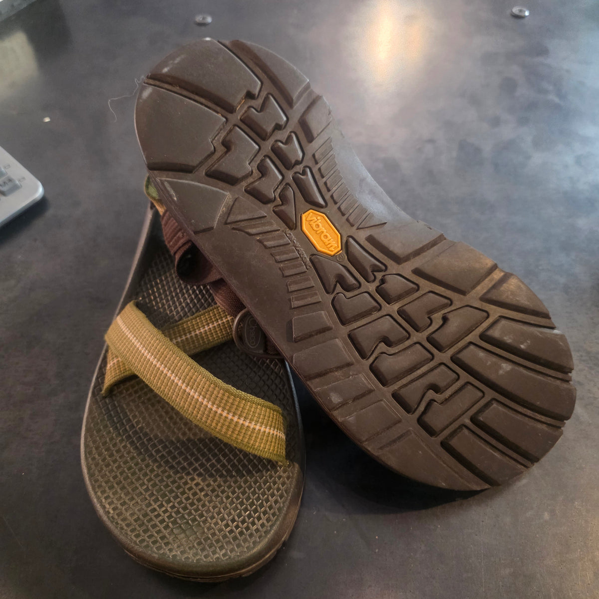 A pair of brown and olive green athletic sandals shown on a dark gray surface, with one sandal turned upside down to display the treaded outsole and a small Vibram logo.