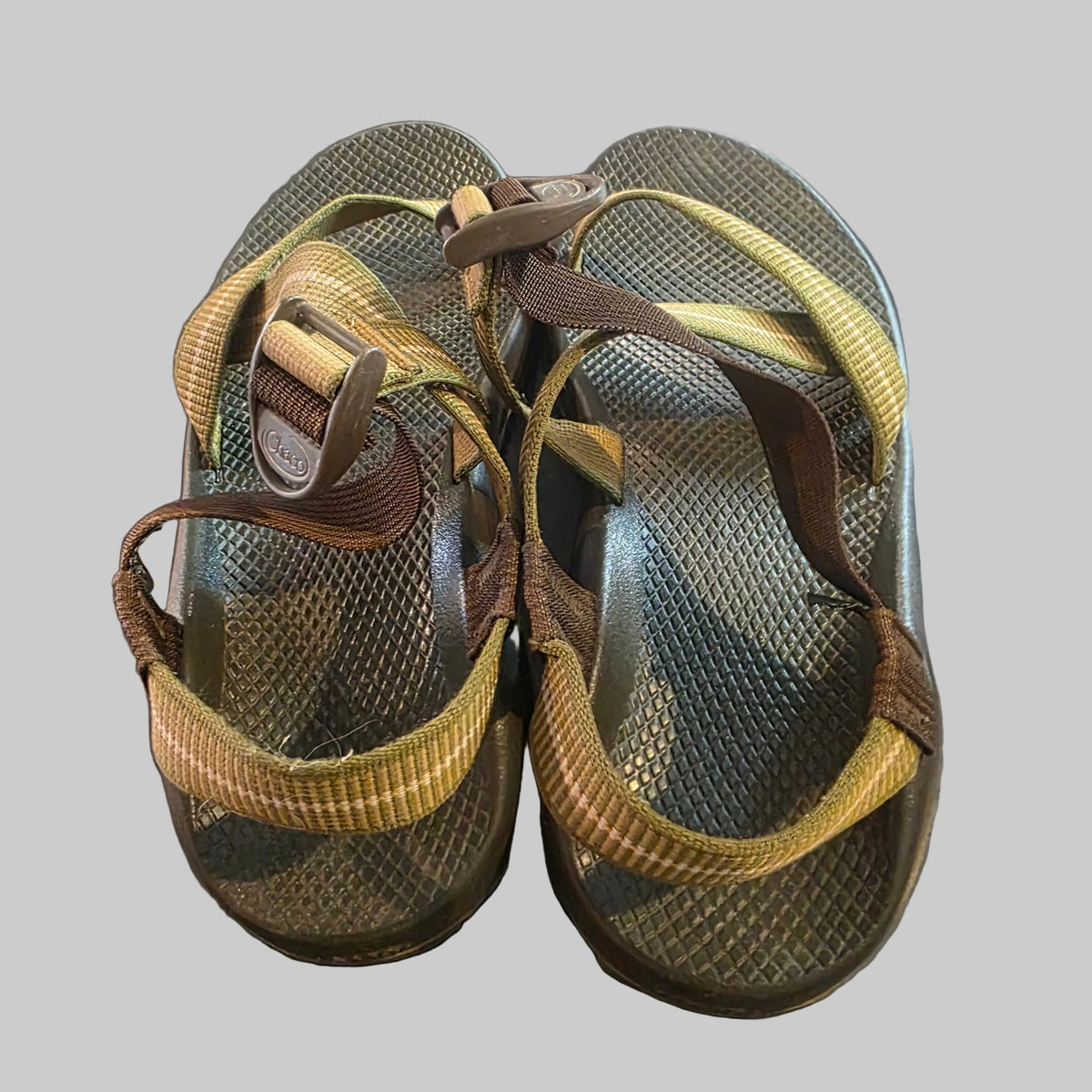A pair of Chaco sandals with olive green mesh footbeds and brown and tan woven straps, shown on a dark gray surface.