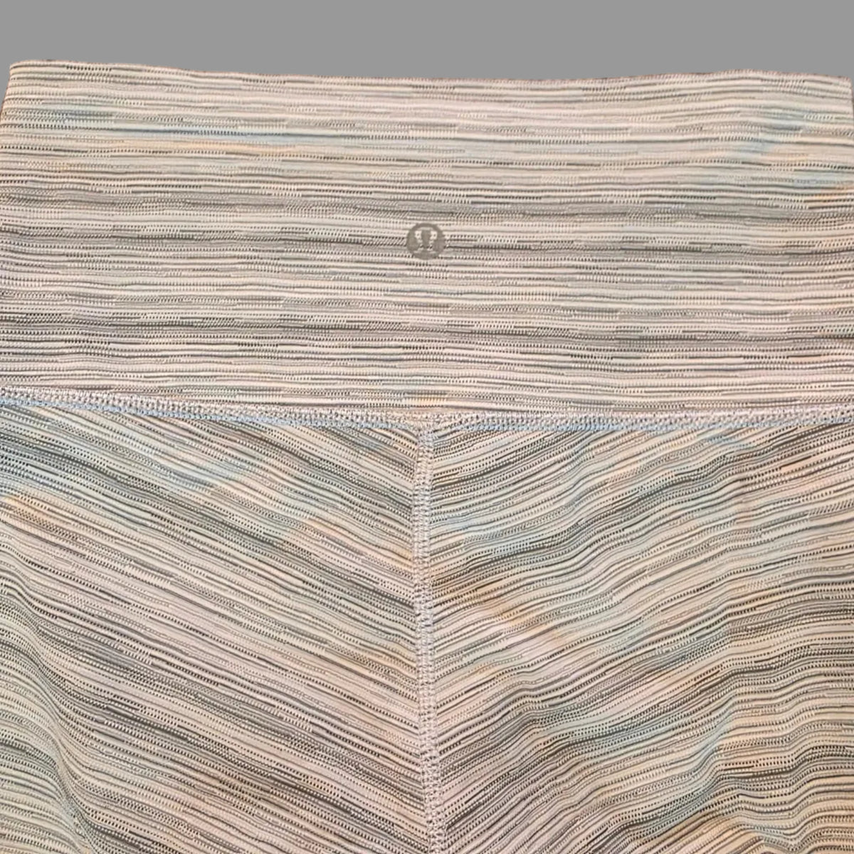 A close-up of a striped textile product laid flat on a speckled brown floor. The fabric has vertical and diagonal stripes in beige, gray, muted blue, and soft green, with visible stitching and a small metal button on the left side.