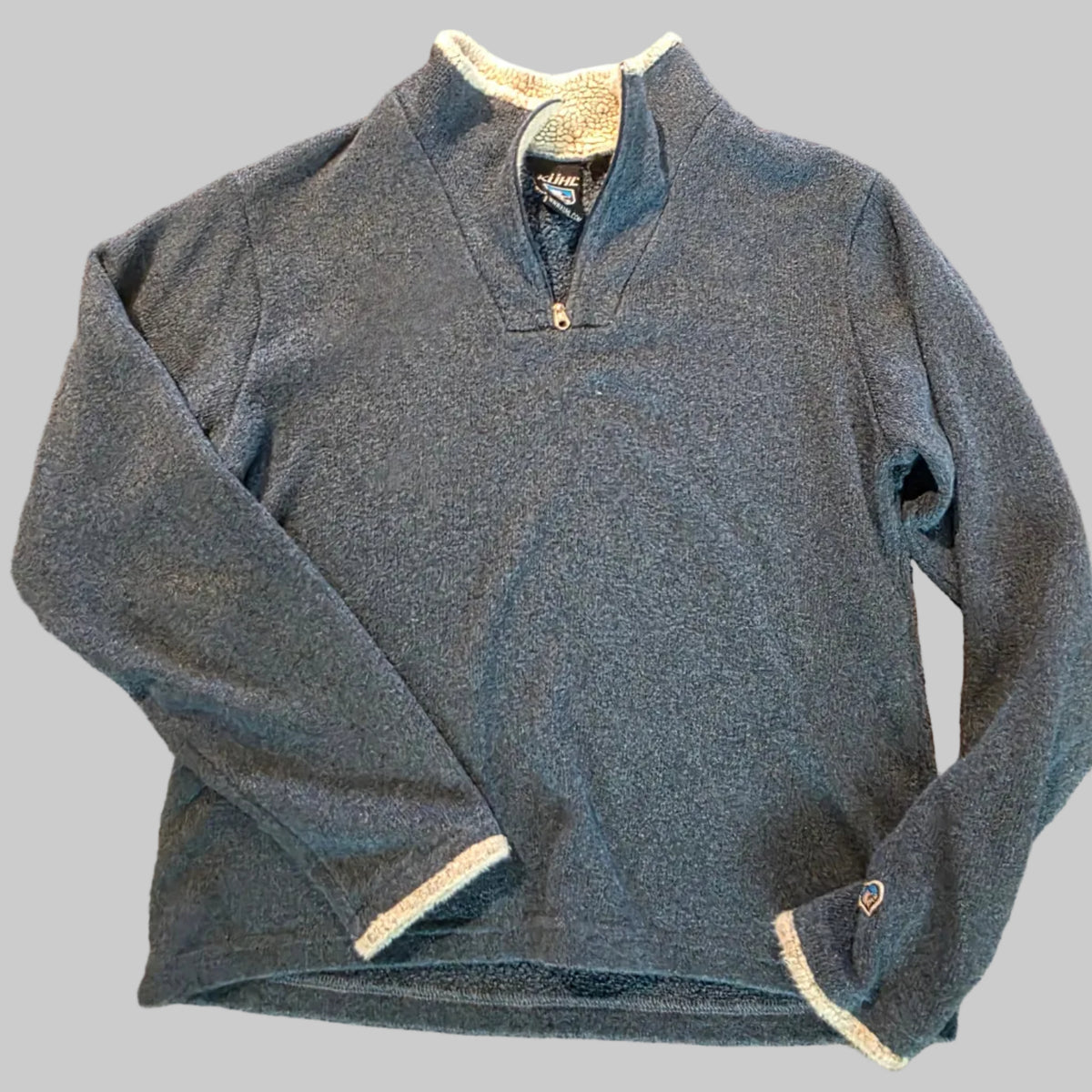A folded heathered blue long-sleeve quarter-zip pullover with cream ribbed trim on the cuffs and collar, laid flat on a textured beige carpet.