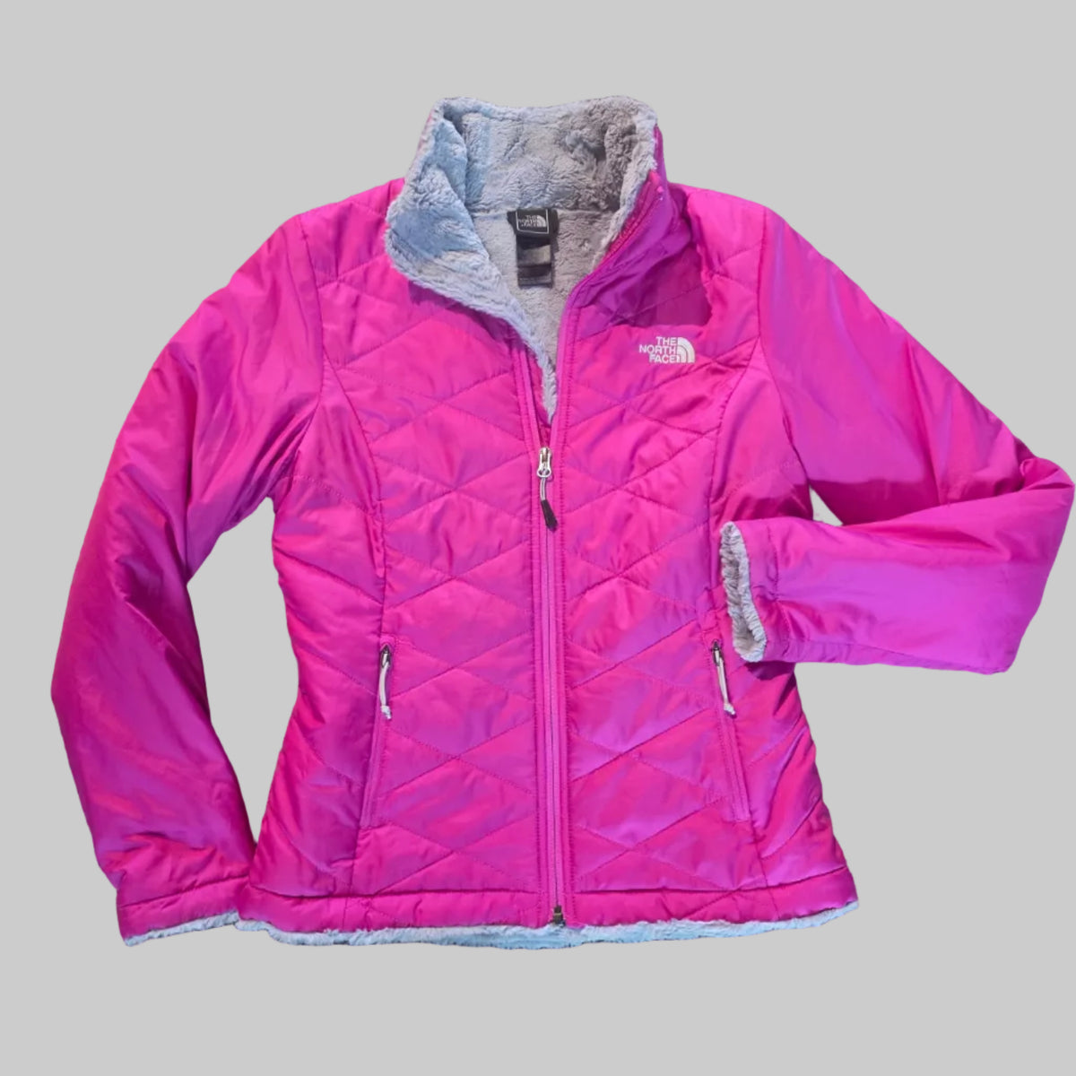 A bright pink quilted puffer jacket with a gray fleece-lined hood and cuffs, laid flat on a brown carpeted floor. The brand logo 'The North Face' is visible on the chest.