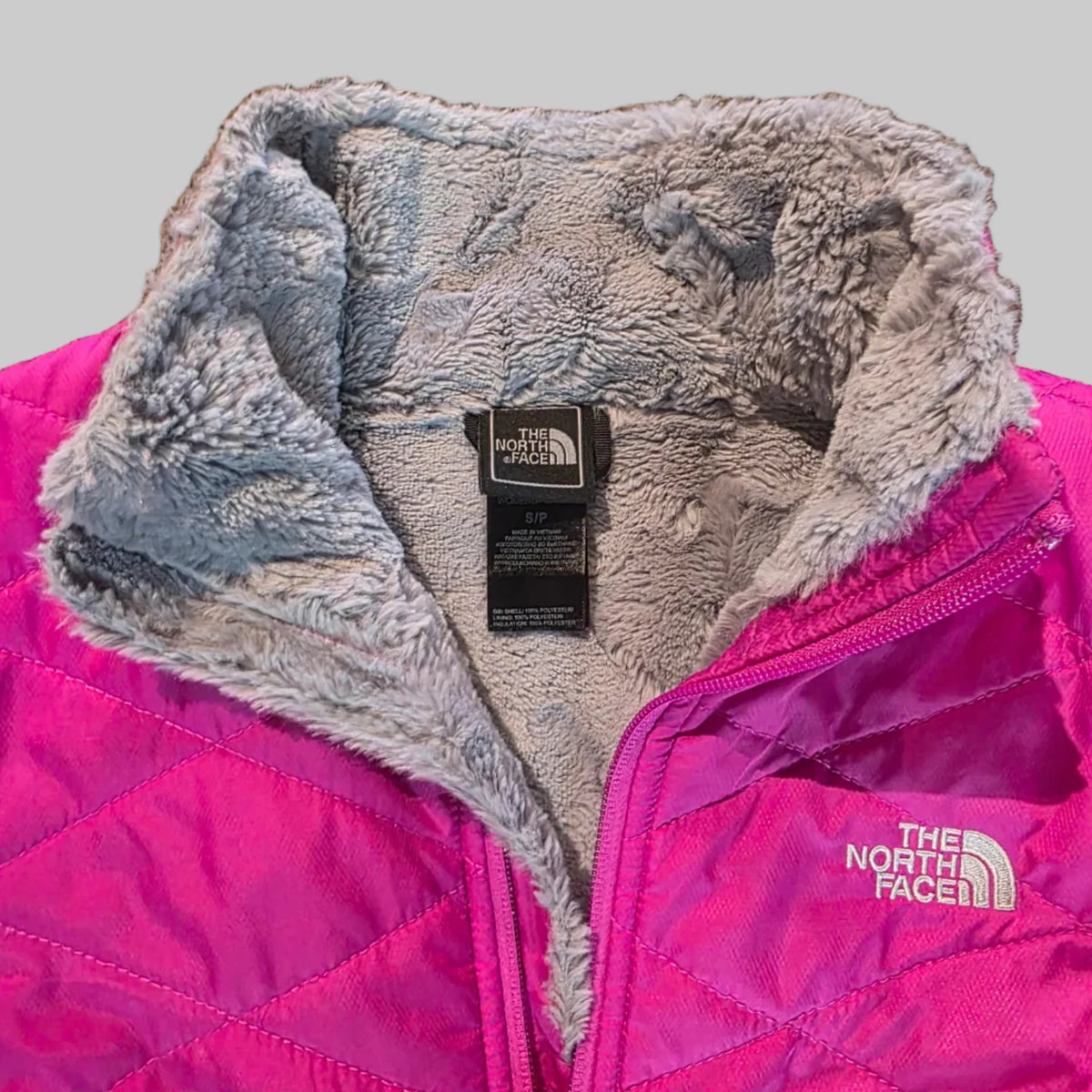 A close-up of a bright pink quilted jacket or sleeping bag with a gray sherpa lining, showing a zippered section, fleece texture, and The North Face brand tag.