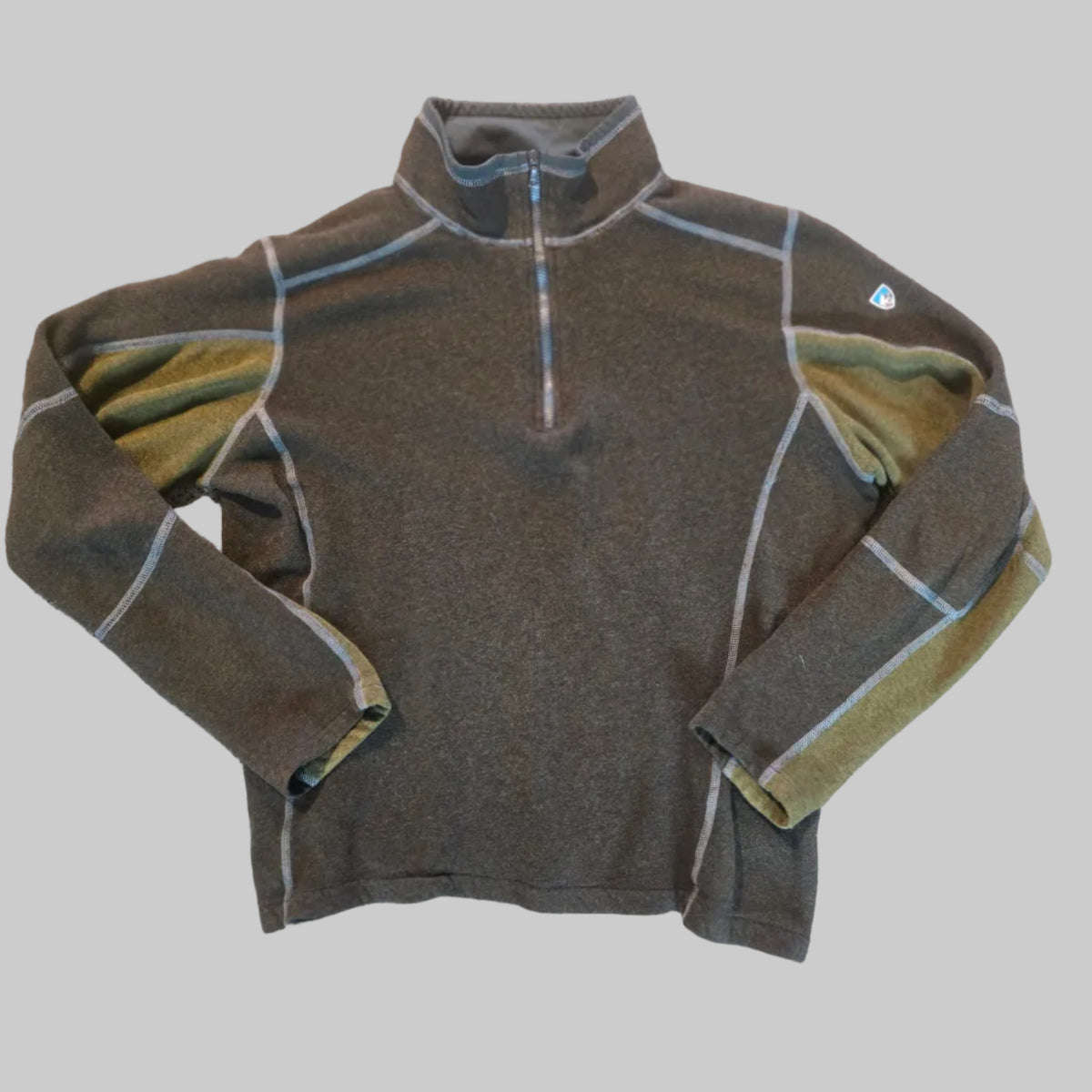 A dark olive brown quarter-zip fleece pullover with contrasting light gray piping and olive green inner lining, laid flat on a textured beige carpet.