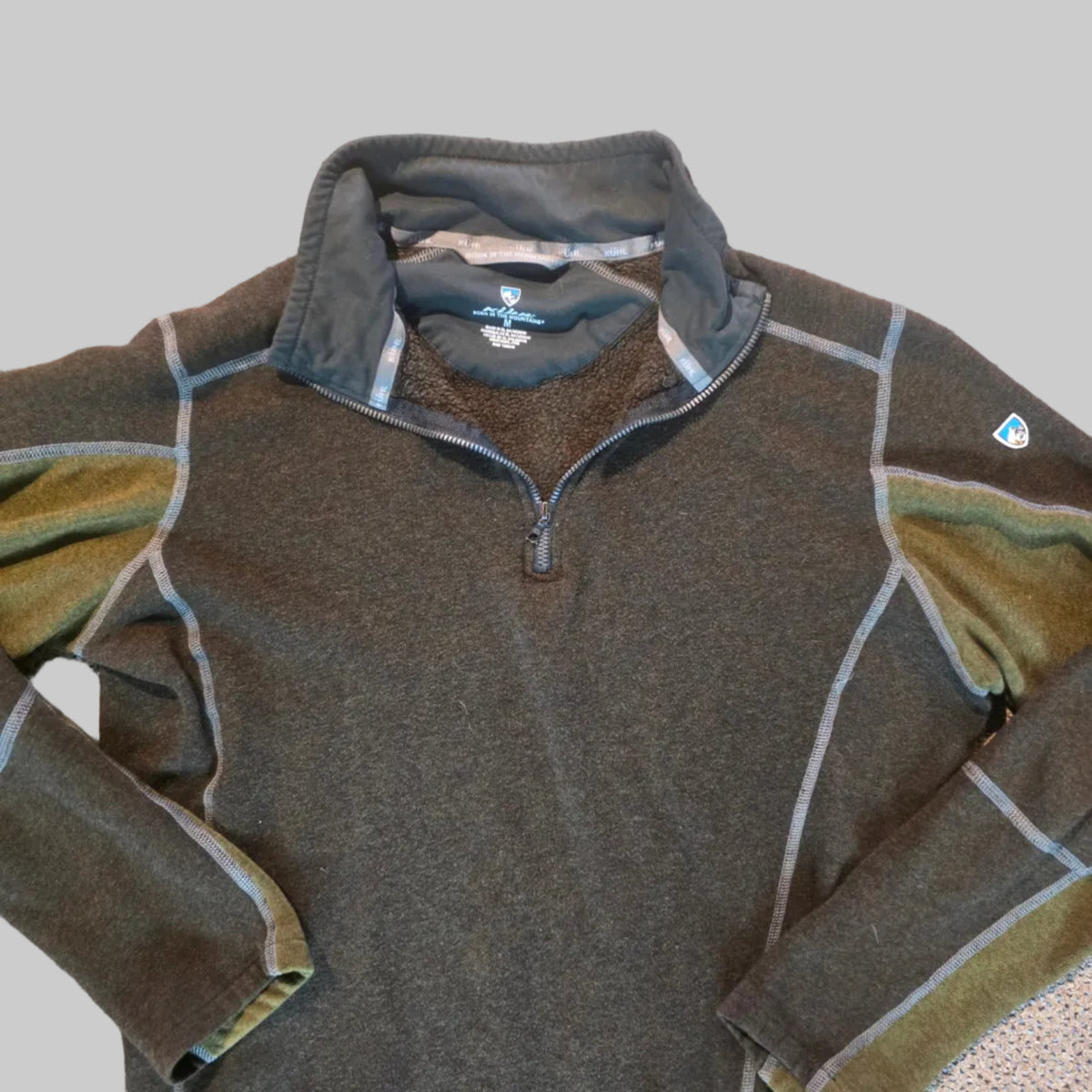 A dark olive green quarter-zip fleece pullover laid flat on a speckled beige carpet. The pullover features contrasting olive green and light gray panels, a half-zip neckline with a gray inner collar, and a small white logo patch near the bottom right hem.