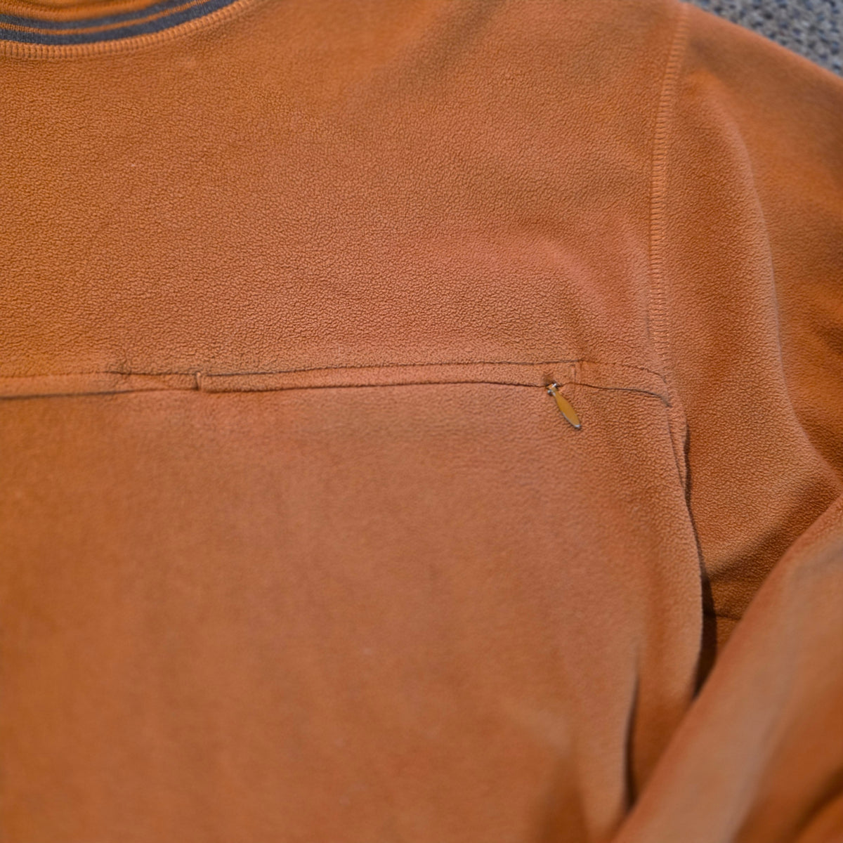 Close-up of a rust-orange fleece pullover sweater showing the chest pocket seam and a small gold zipper pull.