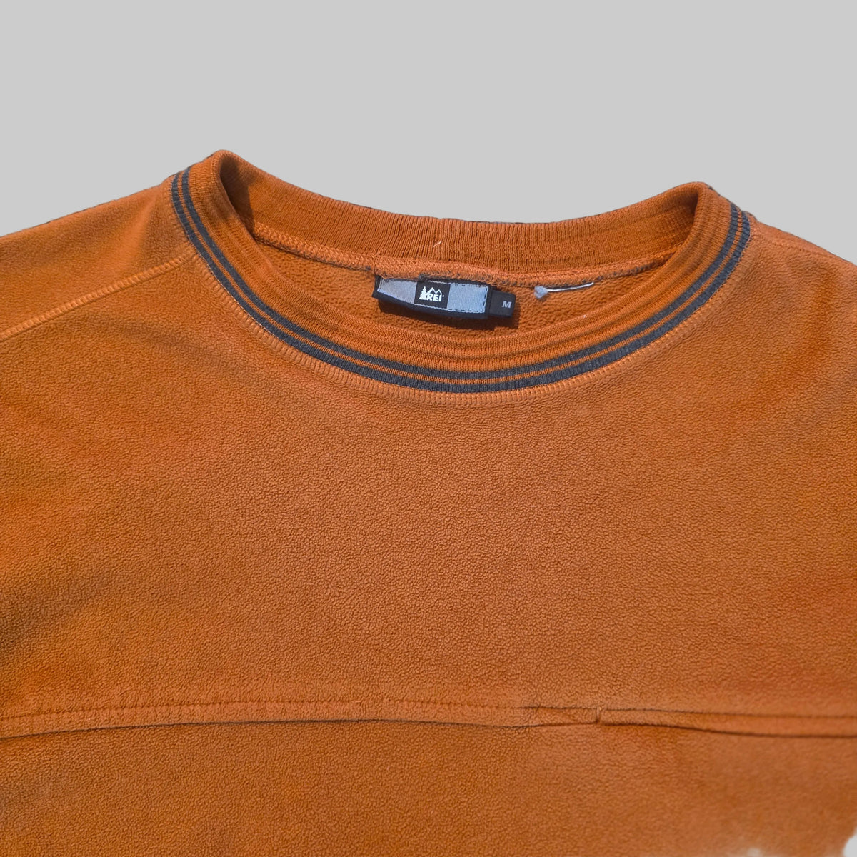 Close-up of an orange crewneck sweatshirt with a dark gray striped ribbed collar, laid flat on a textured gray carpet. The brand name 'A40' is visible on the inner neck tag.