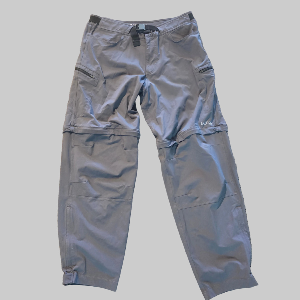 Gray convertible outdoor pants laid flat on a textured beige carpet, featuring zippered pockets and a waistband buckle.