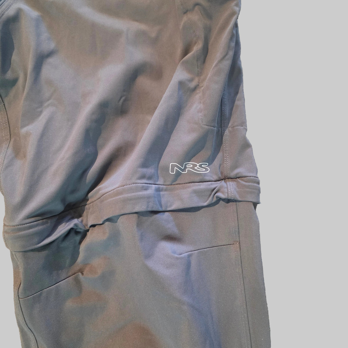 Close-up of gray athletic pants laid flat on a speckled beige floor, showing a zippered pocket and a small white 'NRS' logo near the waistband.