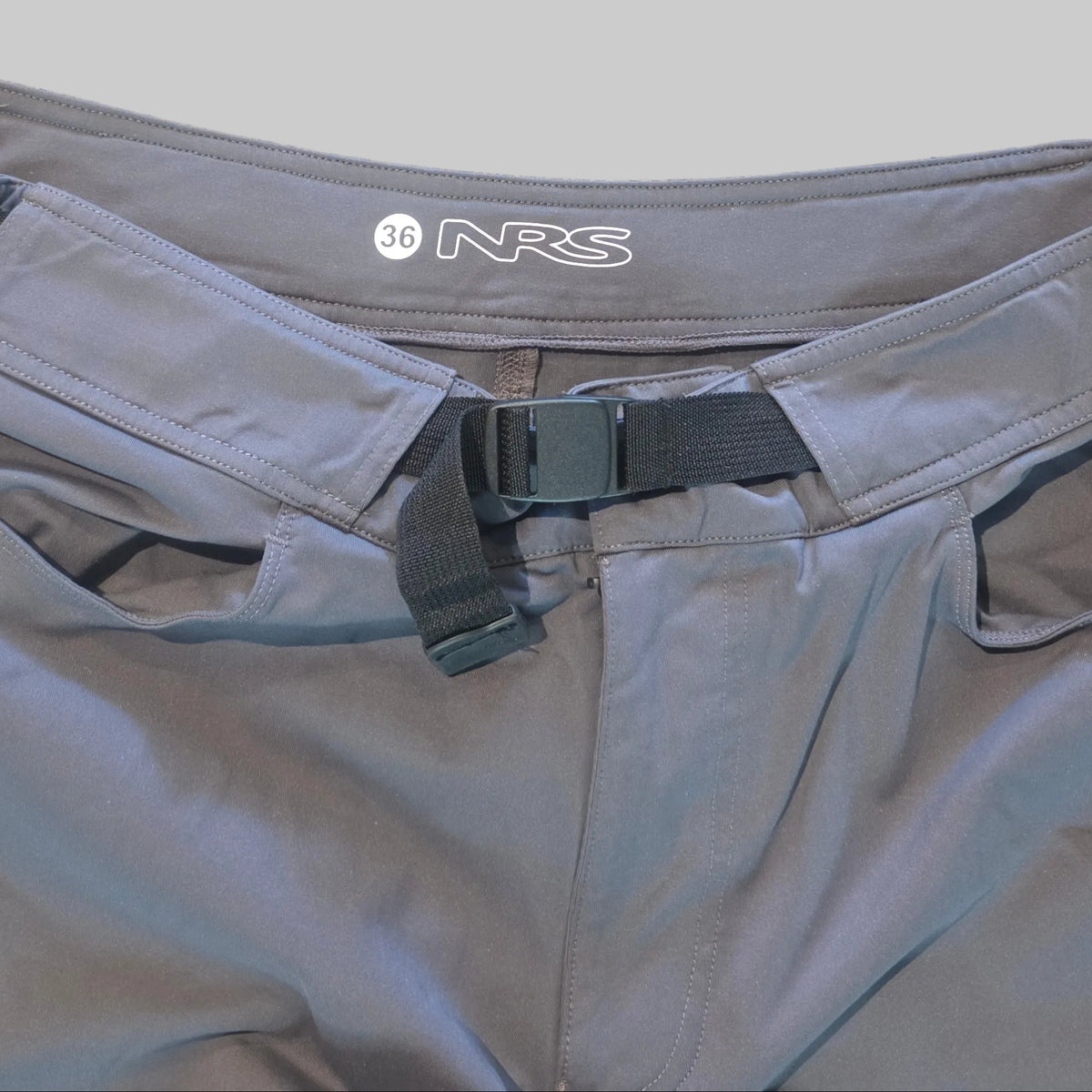 Close-up of gray Snickers Snorkel swim trunks showing the waistband with an adjustable black strap and the white SN logo, laid flat on a speckled beige floor.