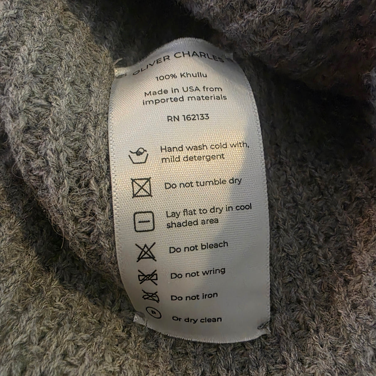 Close-up of a gray knit garment label reading 'OLIVER CHARLES' and care instructions, showing the fabric texture and printed text.