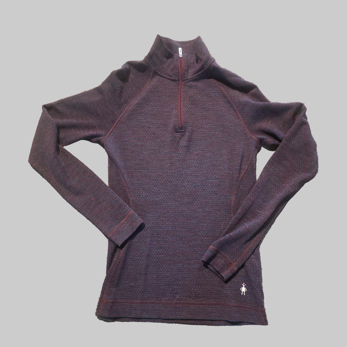 A dark heathered quarter-zip long-sleeve athletic top laid flat on a textured carpet. The garment has a mock neck, raglan sleeves, and a small white polo player logo near the bottom right hem.