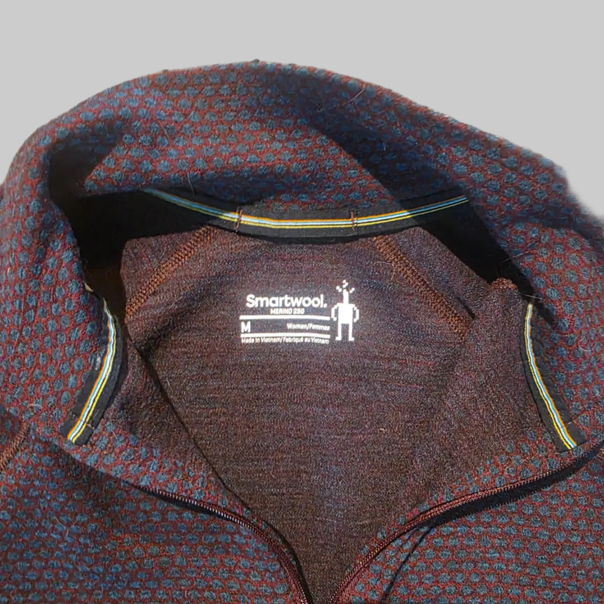 Close-up of the inside neck area of a Smartwool knit garment, showing the white Smartwool logo and a thin yellow and green stripe detail along the collar, set against a textured beige carpet background.