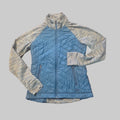 A quilted zip-up jacket laid flat on a textured tan carpet. The jacket has a medium blue body with heathered light gray and blue sleeves and collar.