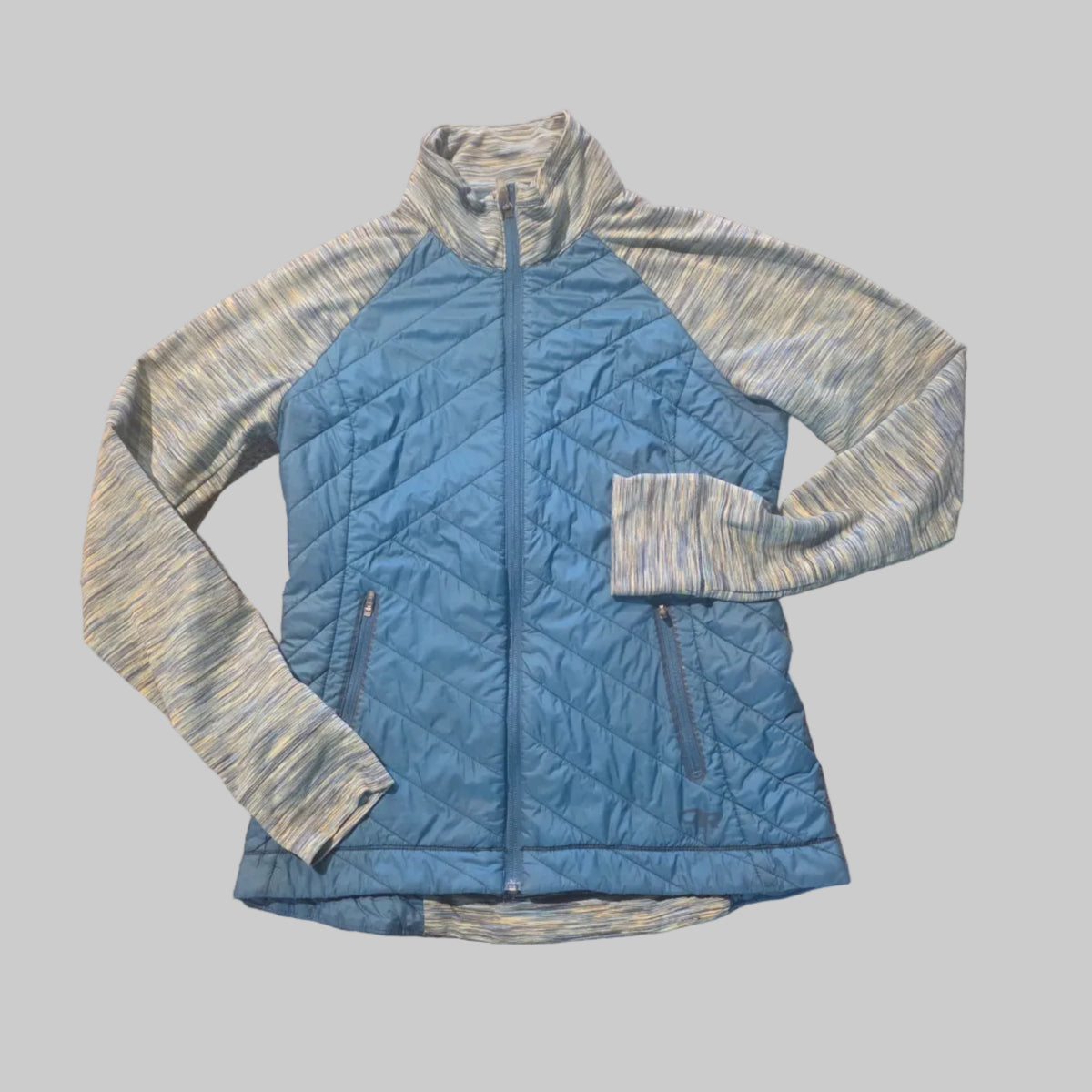 A quilted zip-up jacket laid flat on a textured tan carpet. The jacket has a medium blue body with heathered light gray and blue sleeves and collar.