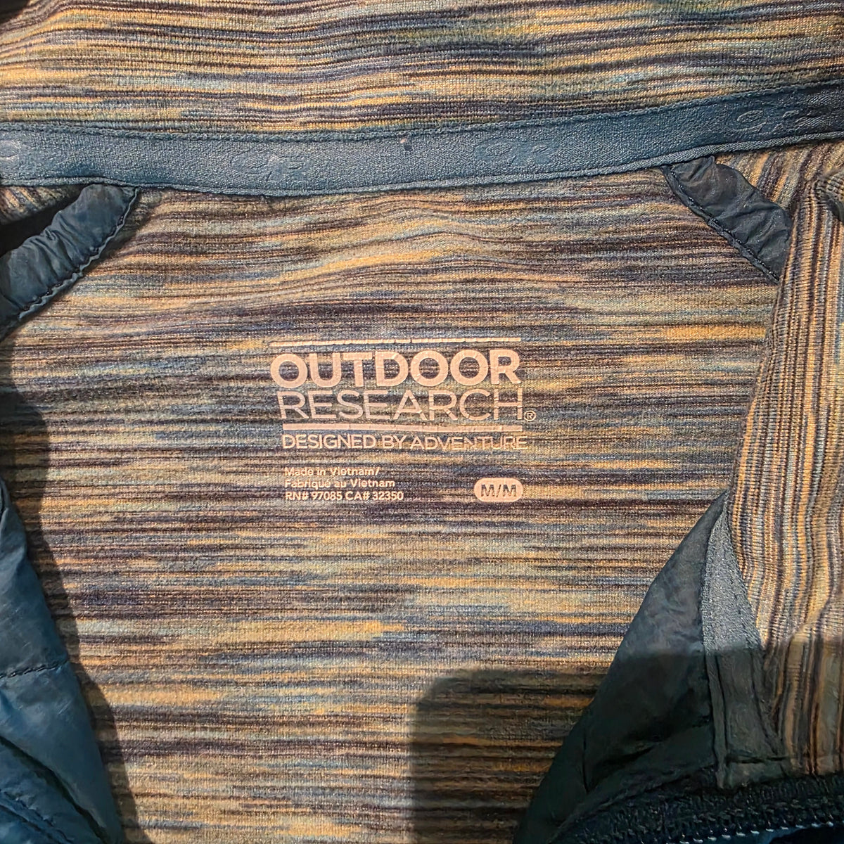 Close-up of a striped technical fabric with vertical lines in brown, tan, and black, featuring a printed label that reads 'OUTDOOR RESEARCH' and 'BEGGED BY ABBEY'.