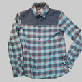 A long-sleeve plaid button-up shirt laid flat on a textured carpet. The shirt features a light blue and teal plaid pattern with thin red stripes, contrasted by solid dark blue panels on the shoulders and collar.