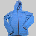 A light blue puffer jacket laid flat on a brown carpeted floor, featuring a central zipper, zippered cuffs, and a small black diamond logo on the chest.