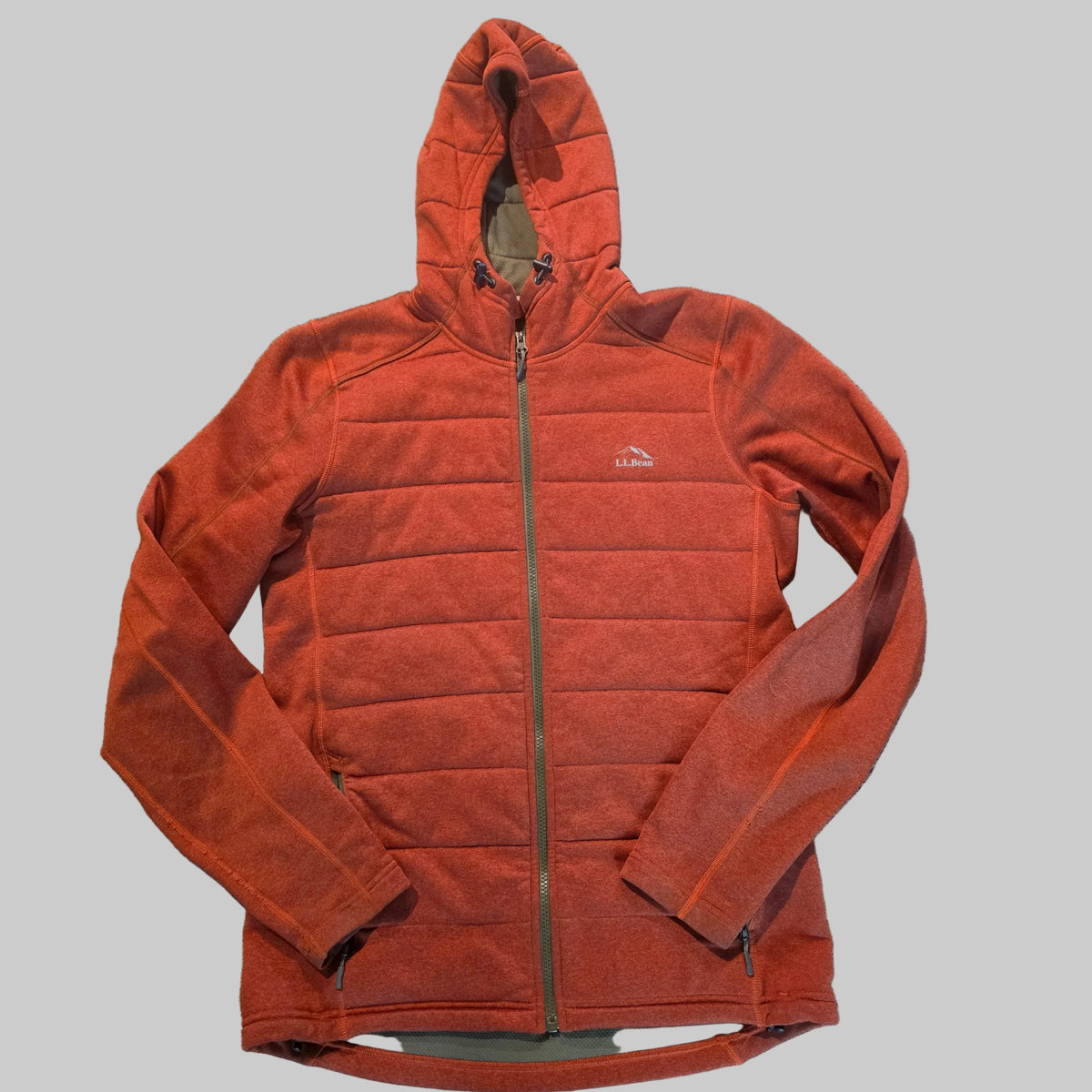 A rust-orange fleece zip-up hoodie with horizontal quilting, laid flat on a gray carpeted floor. The brand logo on the left chest reads 'The North Face'.