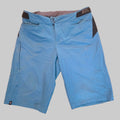 Blue mountain biking shorts with black side panel accents and a gray inner waistband, laid flat on a speckled brown carpet.