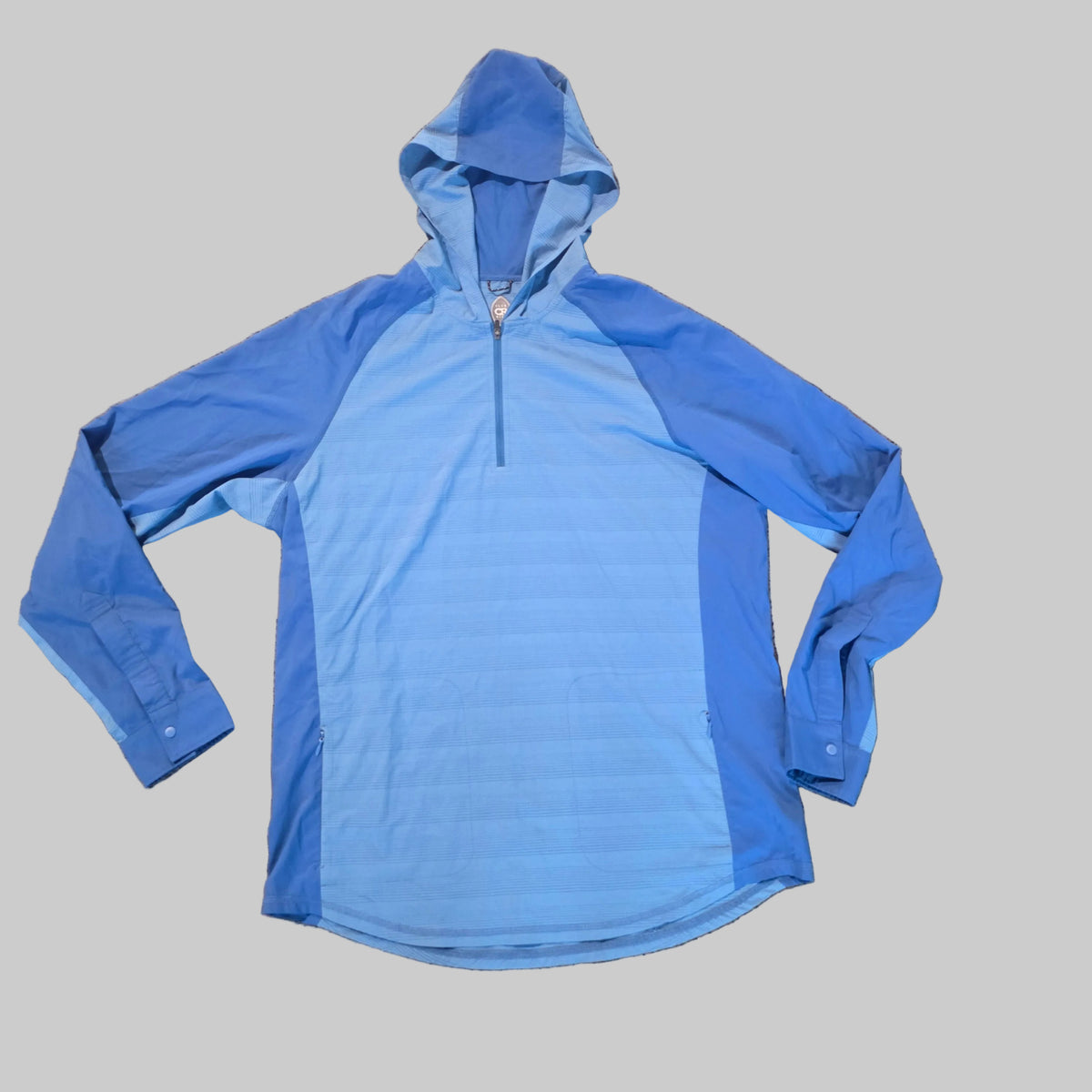 A light blue and medium blue hooded pullover jacket laid flat on a brown carpeted floor.