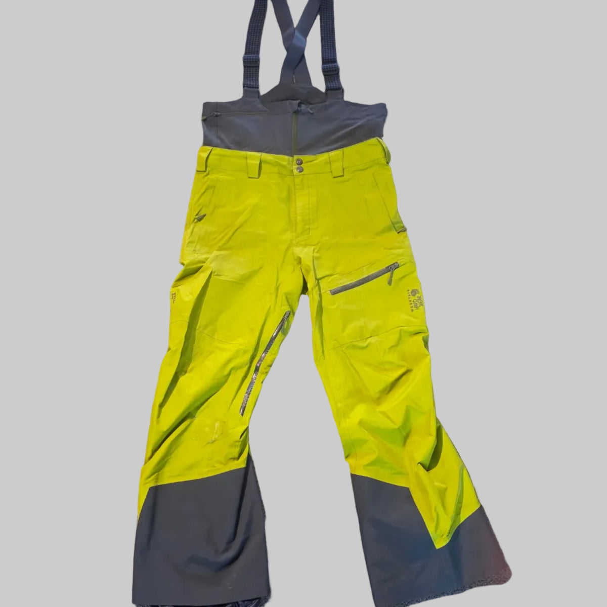 Bright neon yellow and dark gray snow pants with suspenders, laid flat on a gray carpeted floor.