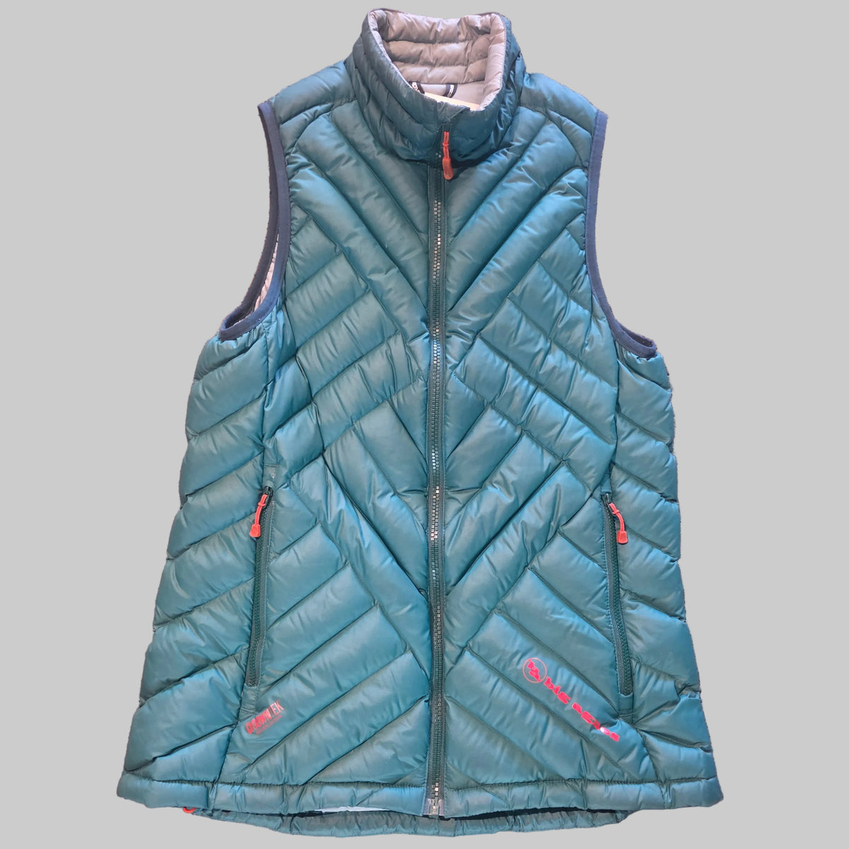 A teal blue diamond-quilted puffer vest with a high stand-up collar, front zipper, and zippered side pockets, laid flat on a textured pinkish-brown carpet. The brand name 'Marmot' is visible in red text near the bottom right hem.