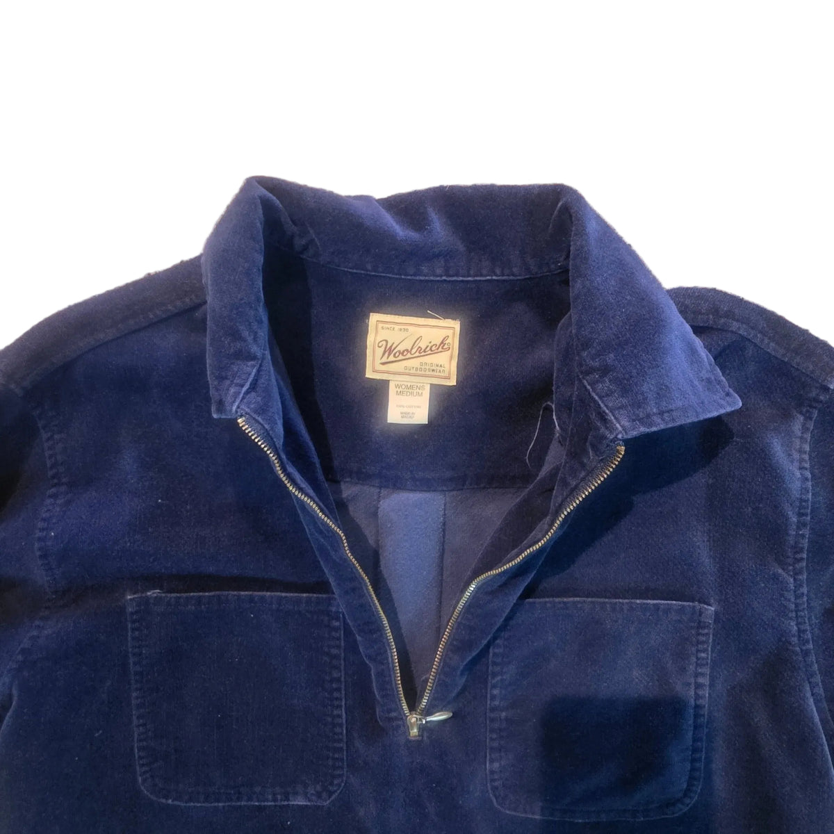 A close-up of a dark blue corduroy jacket laid flat on a carpeted floor, showing the front zipper, two front patch pockets, and an interior brand label.