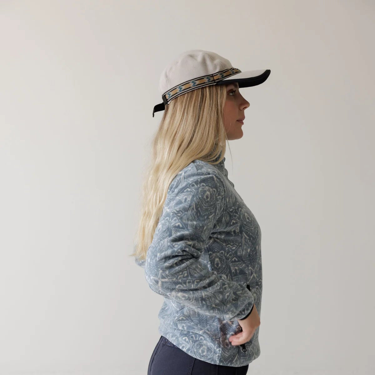 A woman in profile wearing a light blue patterned long-sleeve top, dark navy pants, and a beige cap with a black band, standing against a plain light gray studio background.