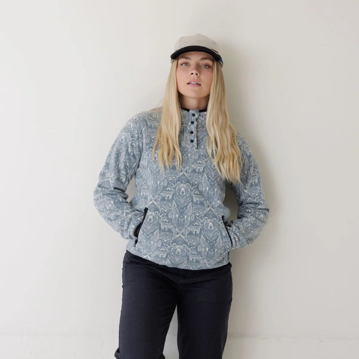 A woman stands against a plain off-white wall wearing a light blue and white patterned quarter-zip pullover, dark navy pants, and a light-colored cap.