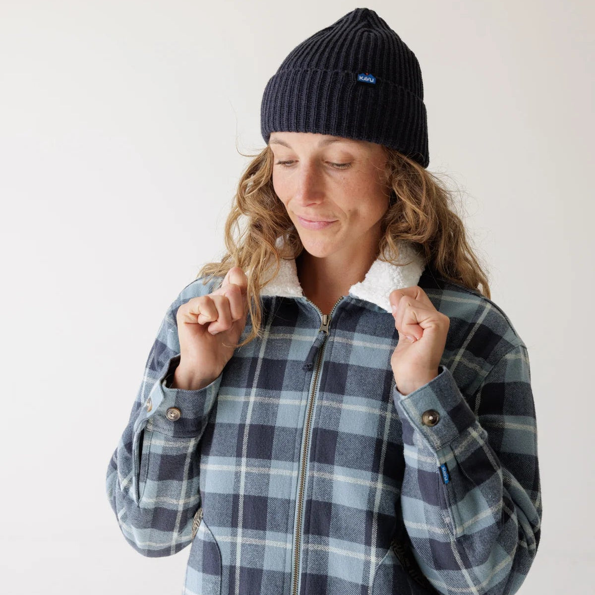 A woman wearing a navy blue and gray plaid zip-up jacket with a white sherpa collar, paired with a dark navy knit beanie and tan pants, posing against a plain off-white studio background.