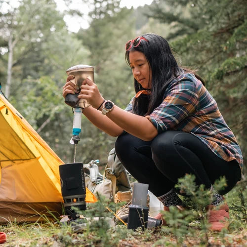Person camping outdoors, using a portable stove near a tent.