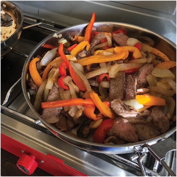A stainless steel pan filled with sliced beef, onions, and colorful bell peppers cooking on a gas stove burner.