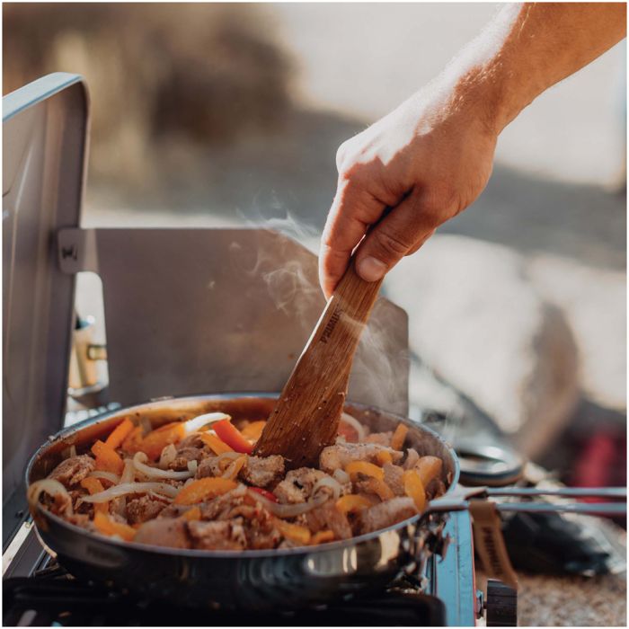 A close-up lifestyle shot of a person's hand using a wooden spatula to stir-fry a pan of meat, peppers, and onions on a portable stove.