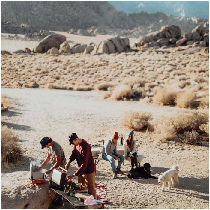 A group of four people camping in a desert landscape with boulders and dry brush. Two people are standing near a small cooking setup with a portable stove and containers, while two others sit in folding chairs nearby.