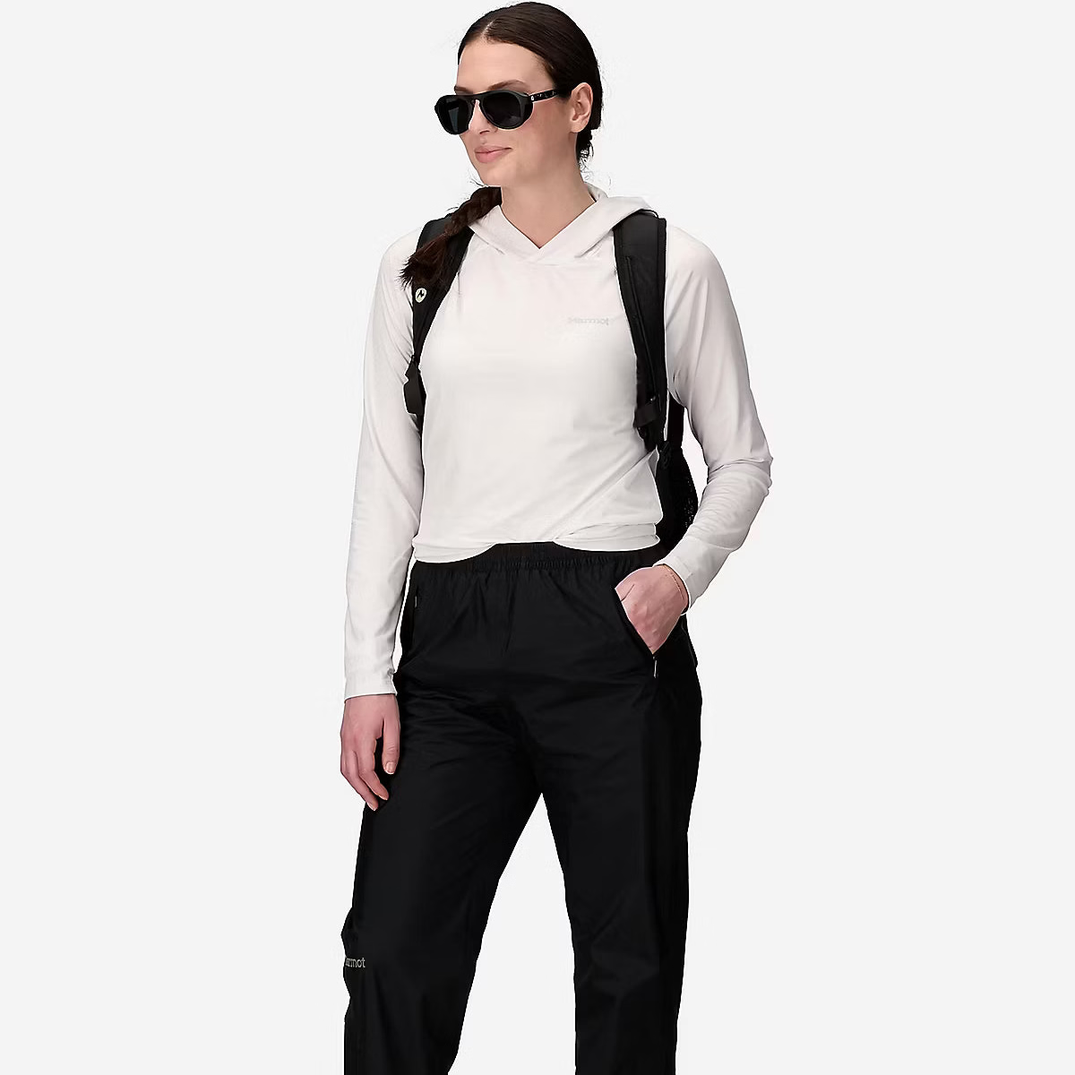 A woman modeling black outdoor pants with a white long-sleeve hooded top, black sunglasses, and a black backpack against a light gray studio background.