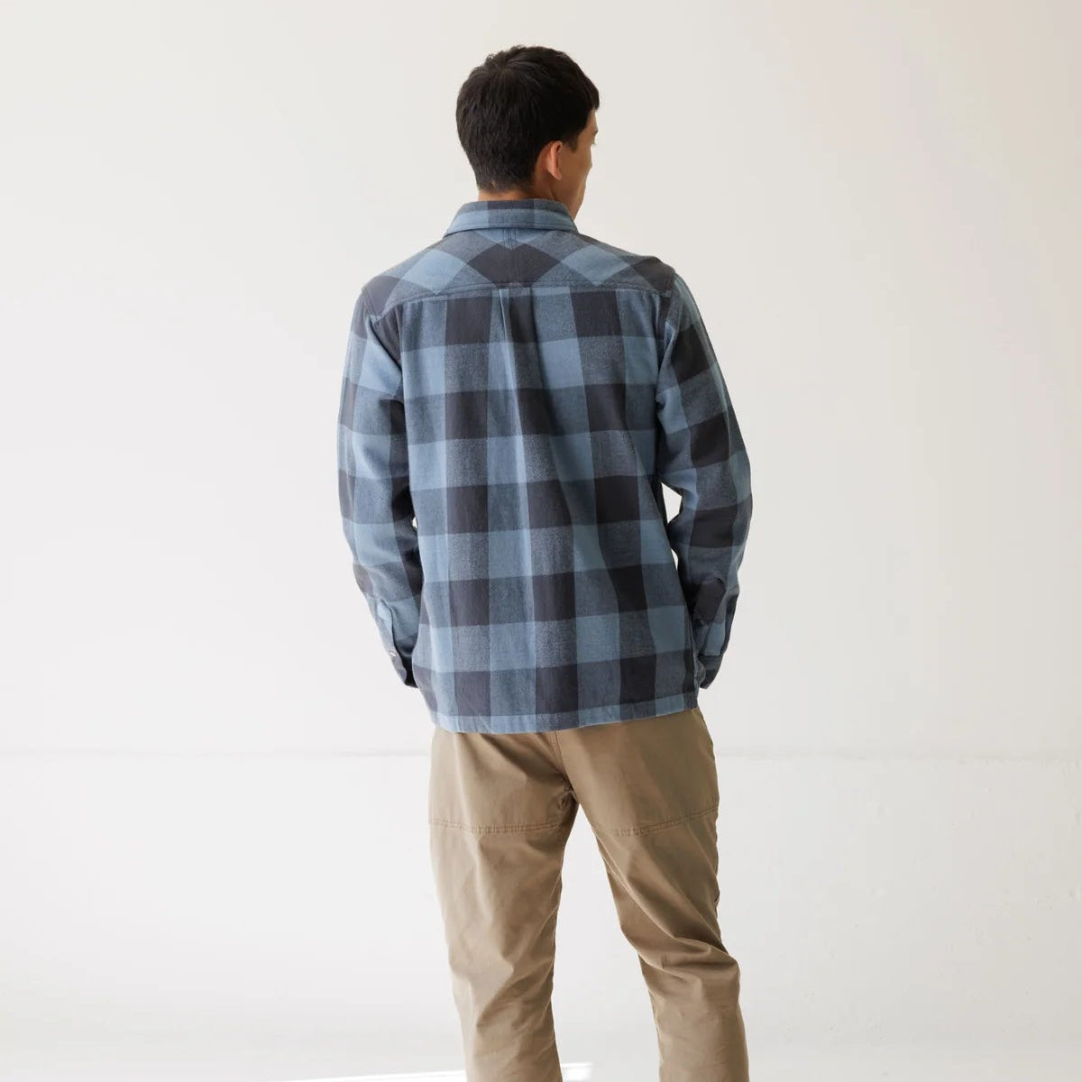 A man viewed from behind wearing a blue and gray plaid flannel shirt, tan pants, and black sneakers, standing in a bright white studio setting.