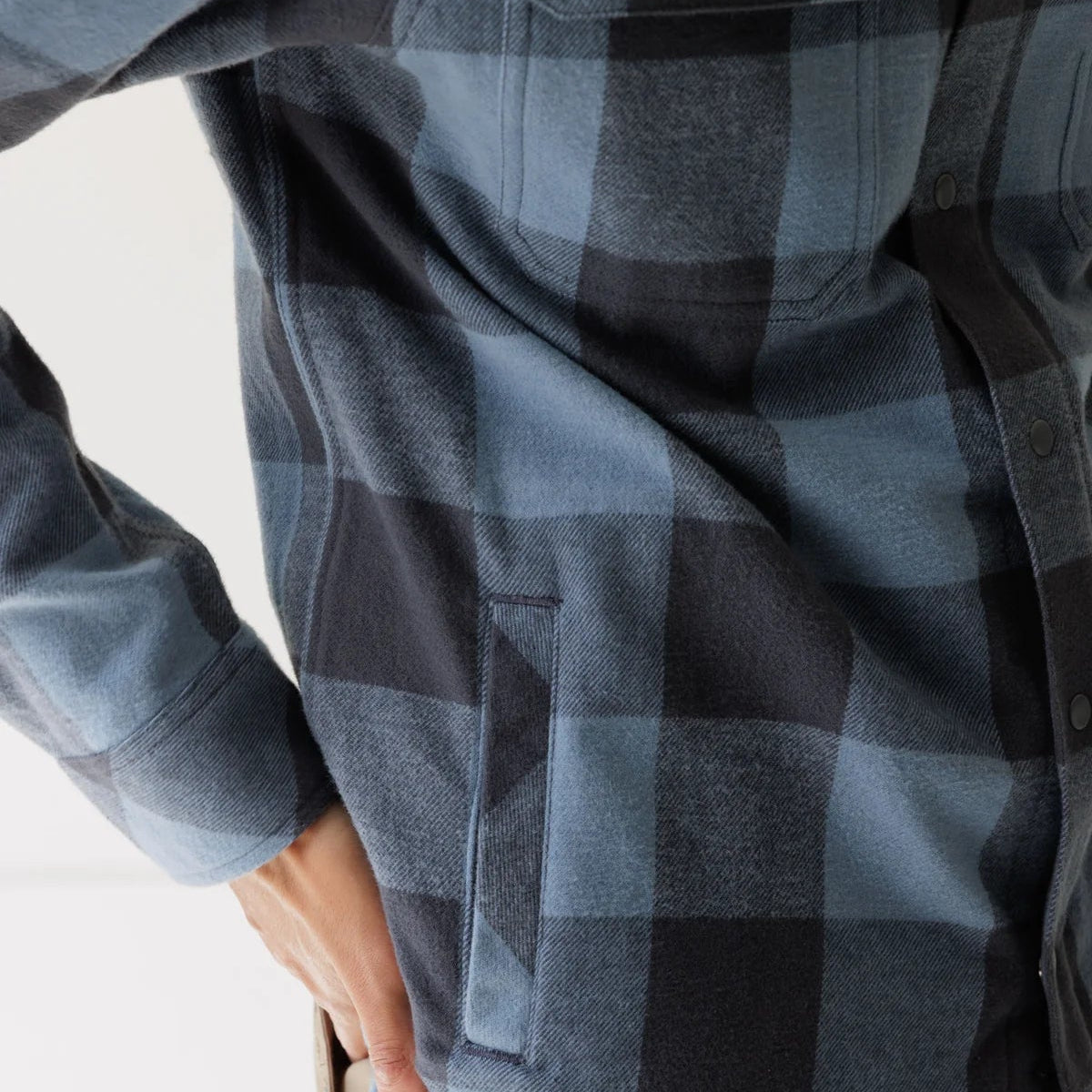Close-up of a person wearing a blue and black buffalo plaid flannel shirt with a side welt pocket, paired with tan pants.