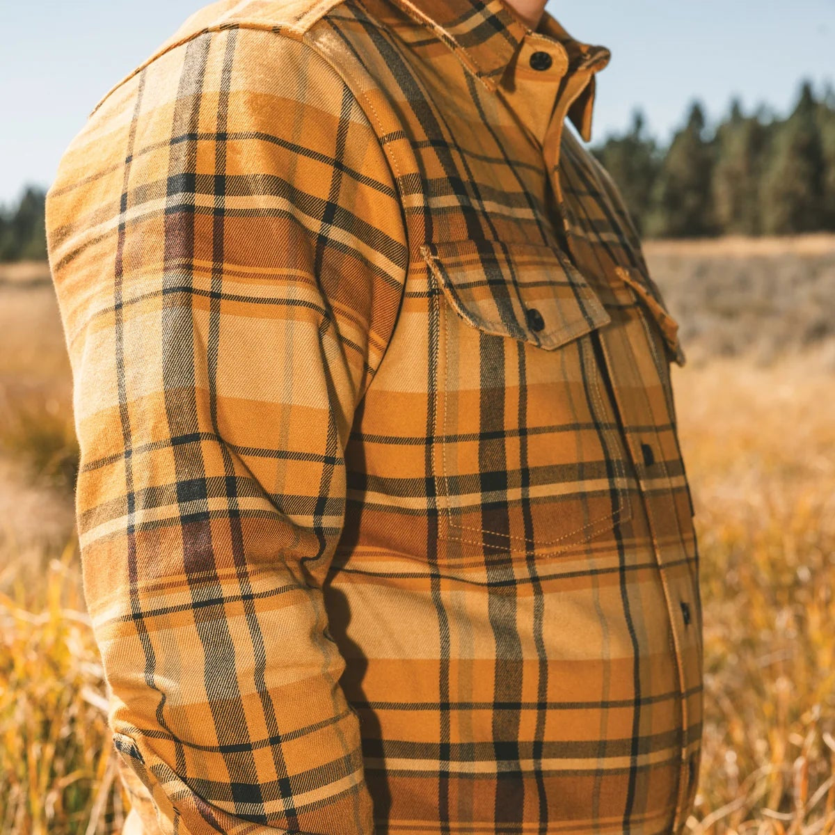 A person standing in a dry grassy field wearing a long-sleeve plaid flannel shirt in mustard yellow, brown, and black, paired with tan pants.