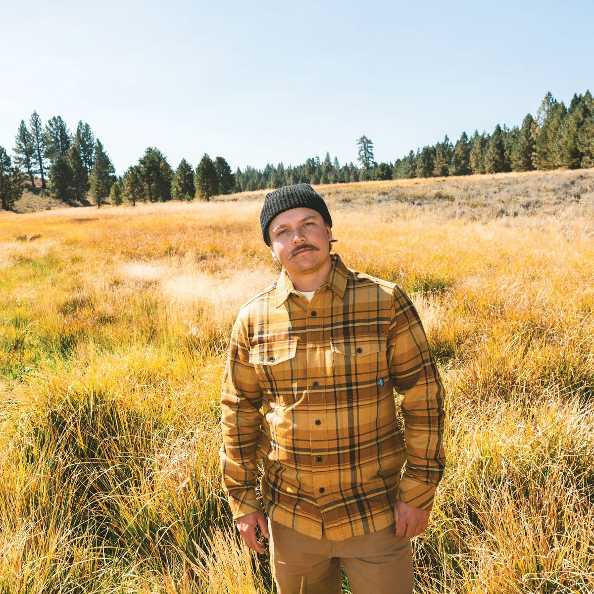 A man wearing a yellow and brown plaid flannel shirt, tan pants, and a black beanie stands in a grassy field under a clear blue sky.