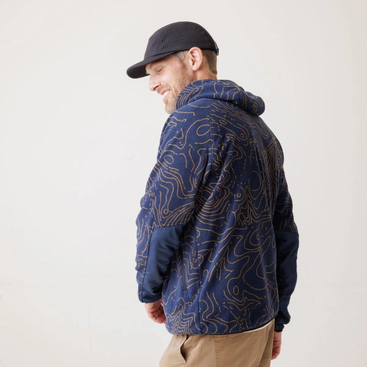 A man wearing a navy blue hooded jacket with a gold topographic contour line pattern, tan pants, and a black baseball cap, photographed against a plain off-white studio background.