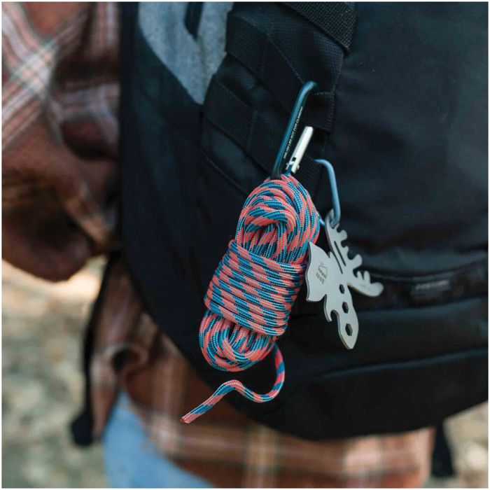 A close-up of a black backpack with a coiled blue and red rope, a teal carabiner, and a silver moose-shaped keychain clipped to the side. A person wearing a brown plaid shirt is partially visible in the background.