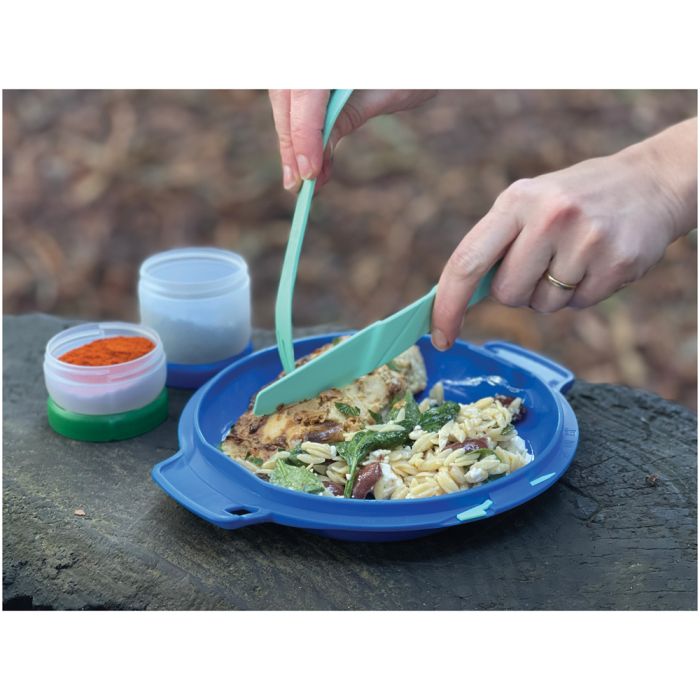 A person uses mint green silicone utensils to eat from a blue fish-shaped plate outdoors on a wooden surface, with small containers of seasoning nearby.