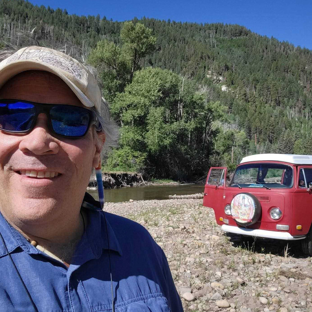 Owner Jeff, with sunglasses and cap standing next to a red vintage van by a river with mountains in the background.