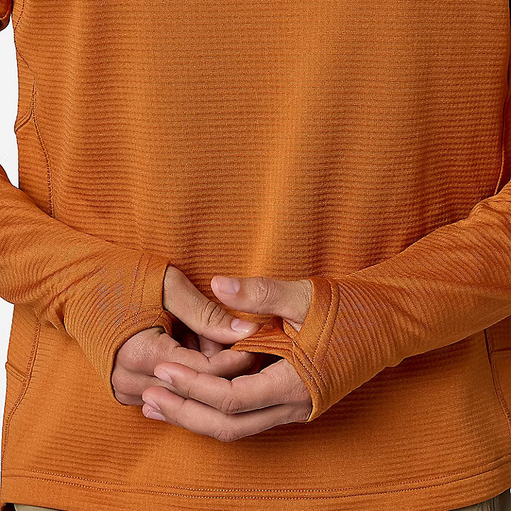 Close-up of a person wearing a textured orange long-sleeve top, with hands folded behind the back and tan pants visible below.